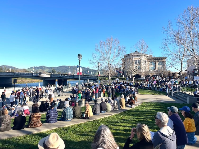crowd of people sitting on concrete rows in a park facing a man at a podium